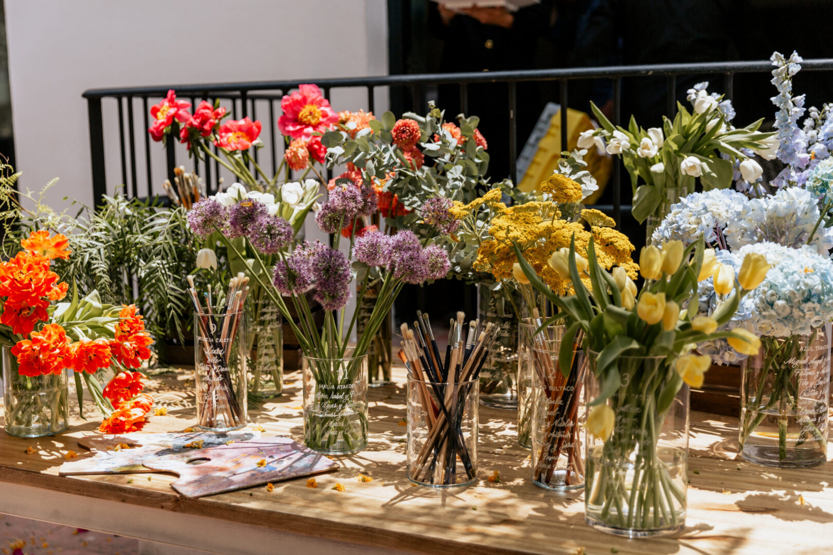Seating plan de boda con nombres grabados en jarrones de cristal con flores de colores vivos junto a paleta de pintor y pinceles sobre mesa de madera