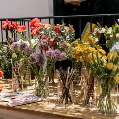 Seating plan de boda con nombres grabados en jarrones de cristal con flores de colores vivos junto a paleta de pintor y pinceles sobre mesa de madera