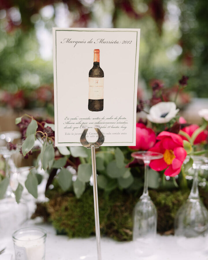Mesero de mesa para boda coleccion Vinos con ilustracion acuarelada de botella de vino tinto sobre soporte alto en mesa de banquete decorada con velas copas de cristal y vajilla elegante