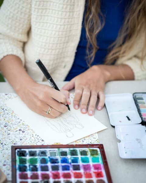 Manos de artista dibujando un boceto a mano con plumilla junto a paletas profesionales de acuarela durante evento de live art