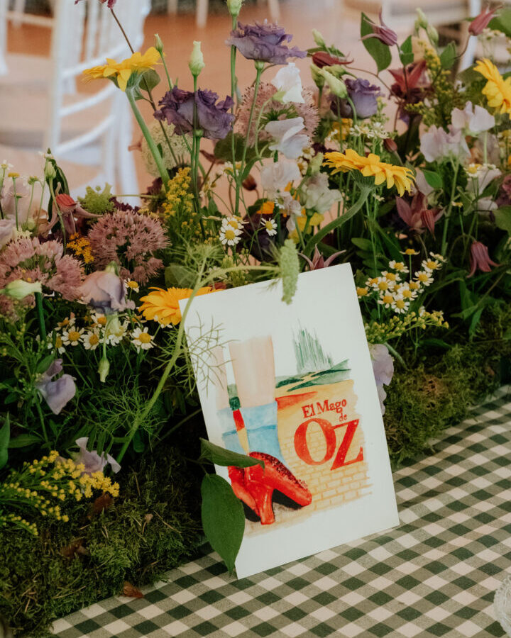 Mesero de boda personalizado con ilustración en acuarela de El Mago de Oz sobre mesa decorada con flores naturales y vegetación