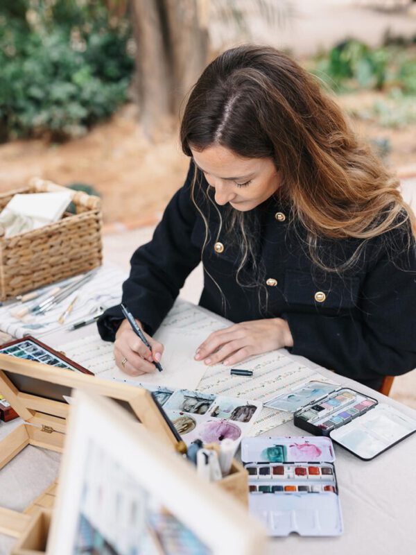 Artista de acuarela pintando en directo en un evento al aire libre con paletas de colores y pinceles sobre mesa de trabajo