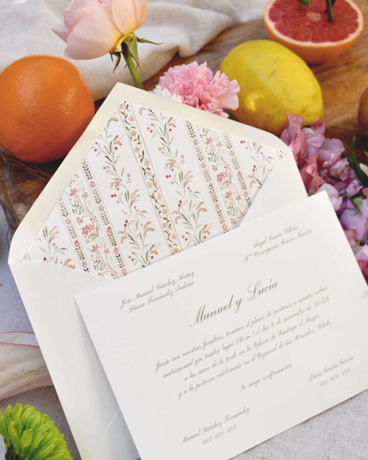 Invitacion de boda coleccion Vintage con cenefa floral rosa y caligrafia manuscrita presentada en composicion lifestyle con citricos hierbas aromaticas y sobre artesanal coordinado