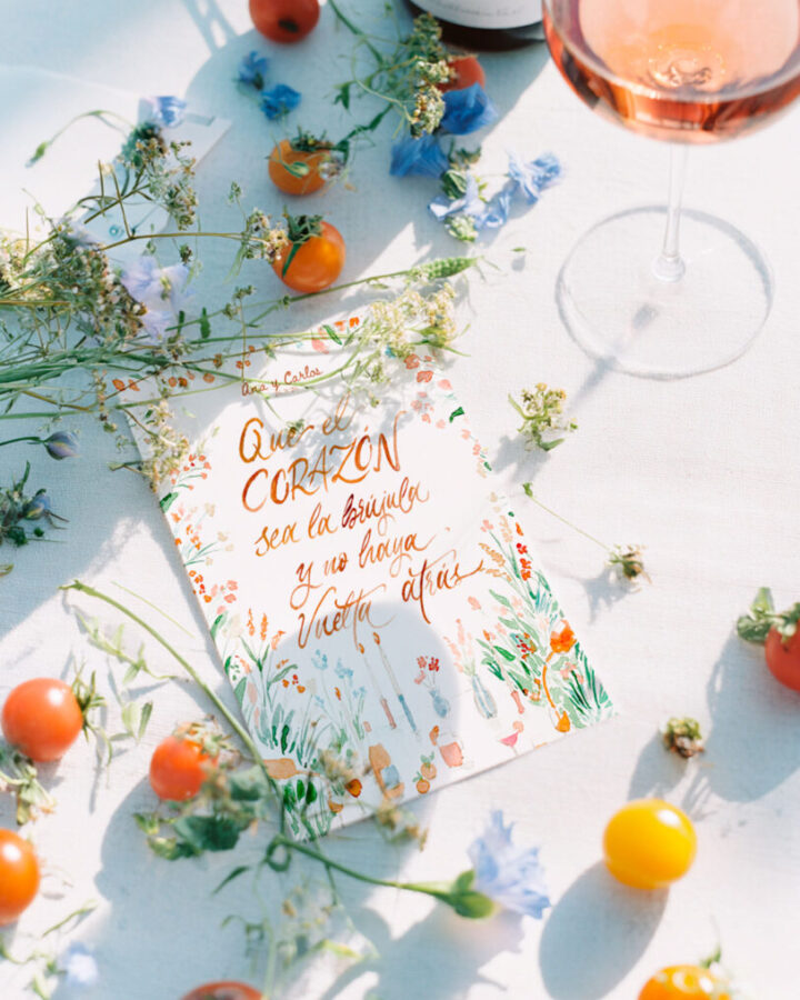 Lámina Bloom con frase en caligrafía artística presentada sobre mesa decorada con flores silvestres, frutas y copas de vino rosado