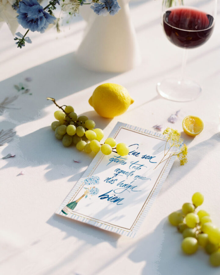 Lámina con frase en caligrafía artística colección Hortensia presentada sobre mesa decorada con flores, frutas y copa de vino