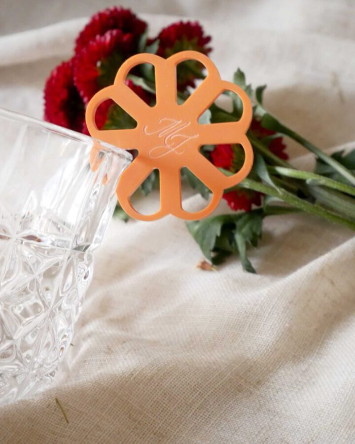 Marcacopas modelo Frida en metacrilato naranja con iniciales grabadas y borde festoneado colgado en copa de cristal para boda