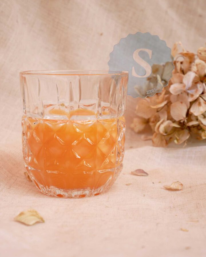 Marcacopas de metacrilato con inicial caligráfica colocado en copa de cóctel con fondo cálido y decoración floral para evento nupcial