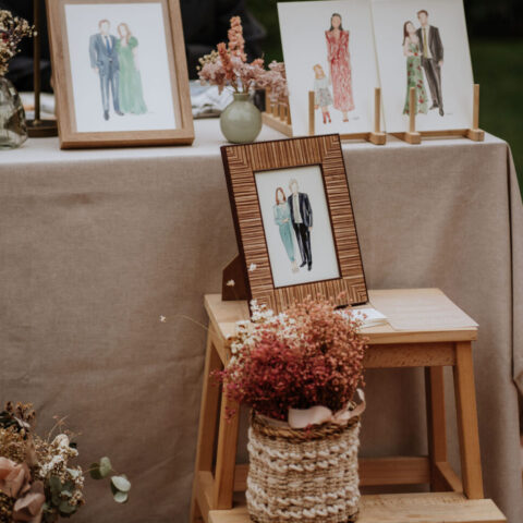 Retratos en acuarela de parejas e invitados expuestos en marcos y caballetes sobre mesa decorada con flores secas en boda