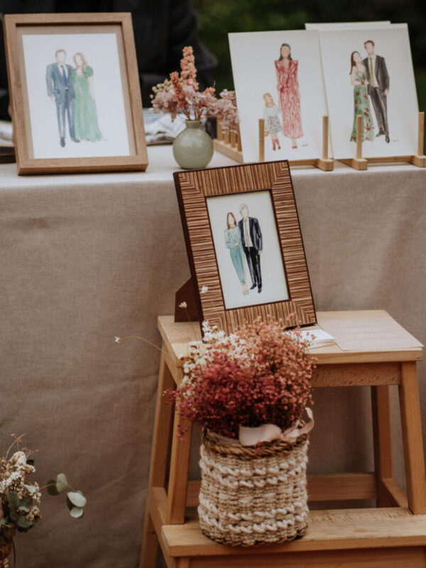 Retratos en acuarela de parejas e invitados expuestos en marcos y caballetes sobre mesa decorada con flores secas en boda