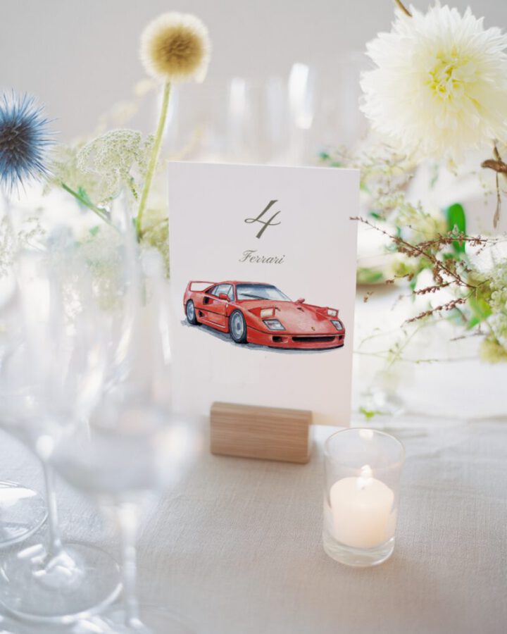 Número de mesa para boda con ilustración en acuarela de Ferrari rojo sobre soporte elegante en ambiente nupcial con velas