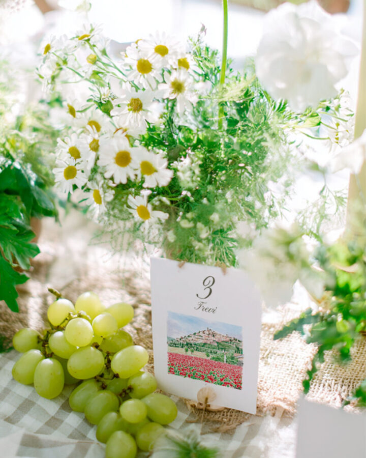 Mesero de boda con número de mesa e ilustración en acuarela de pueblo con paisaje, presentado sobre soporte decorativo con flores