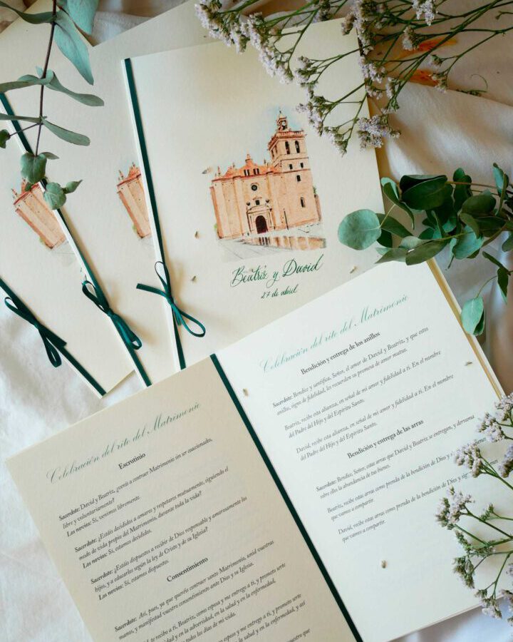Misal de boda artesanal abierto mostrando portada con ilustración en acuarela de iglesia y páginas interiores con textos litúrgicos maquetados