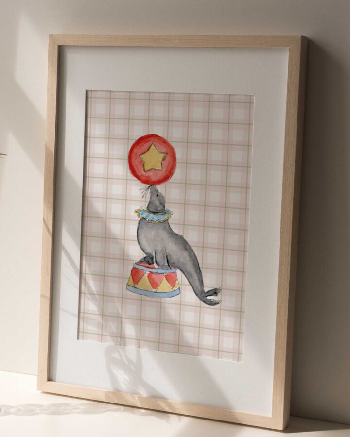 Lámina enmarcada con ilustración de foca de circo pintada en acuarela equilibrando una pelota roja con estrella sobre pedestal