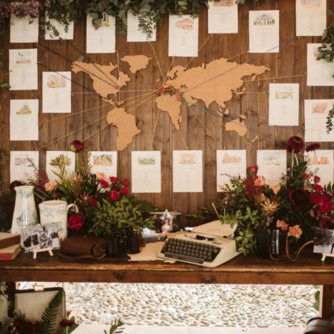Seating plan de boda con mapamundi de madera, meseros ilustrados en acuarela con destinos del mundo, flores rojas y máquina de escribir vintage
