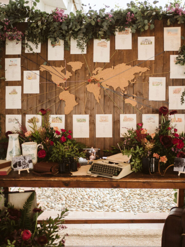 Seating plan de boda con mapamundi de madera, meseros ilustrados en acuarela con destinos del mundo, flores rojas y máquina de escribir vintage