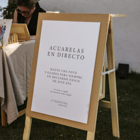 Caballete de madera con cartel de acuarelas en directo de Trantán Estudio en evento al aire libre con texto informativo para invitados