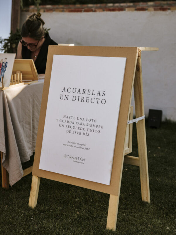 Caballete de madera con cartel de acuarelas en directo de Trantán Estudio en evento al aire libre con texto informativo para invitados