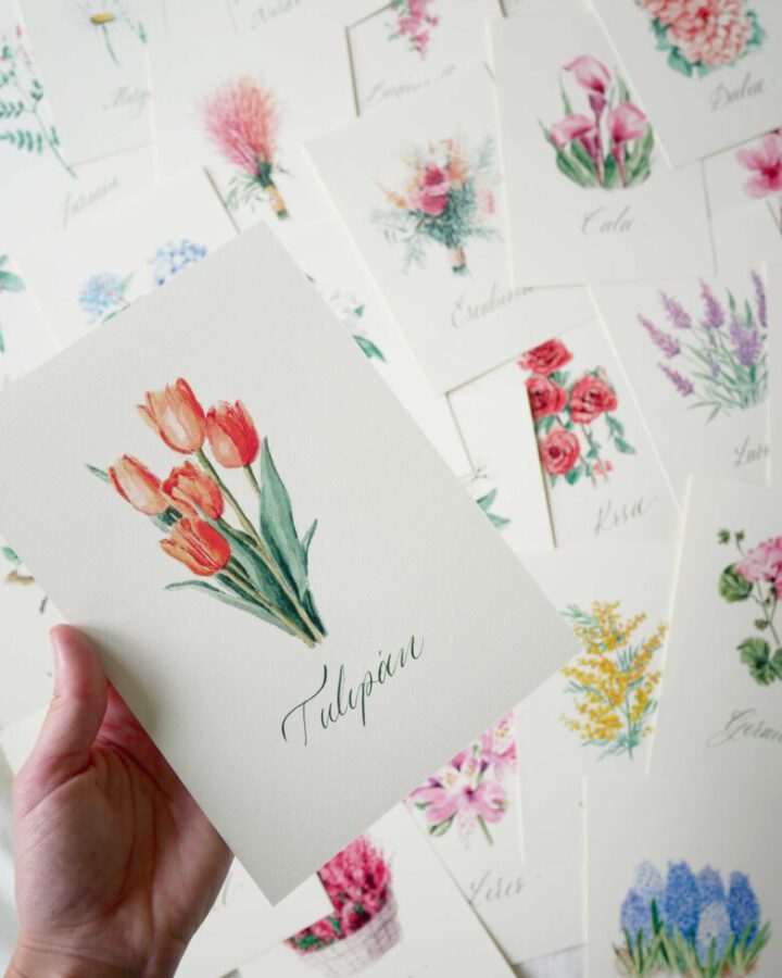 Mano sosteniendo abanico de tarjetas de mesero de boda de la coleccion Flores con ilustraciones de tulipan lavanda y otras flores silvestres pintadas a mano en acuarela artesanal