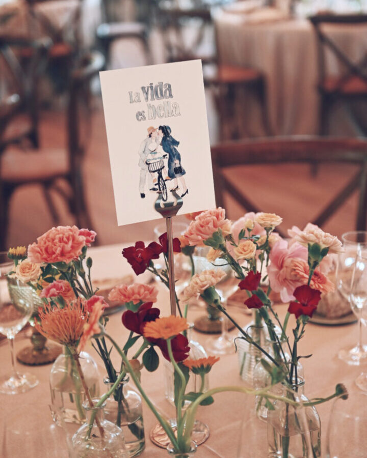 Mesero de mesa con ilustración en acuarela de La Vida es Bella sobre soporte decorativo con flores y ambientación de boda elegante