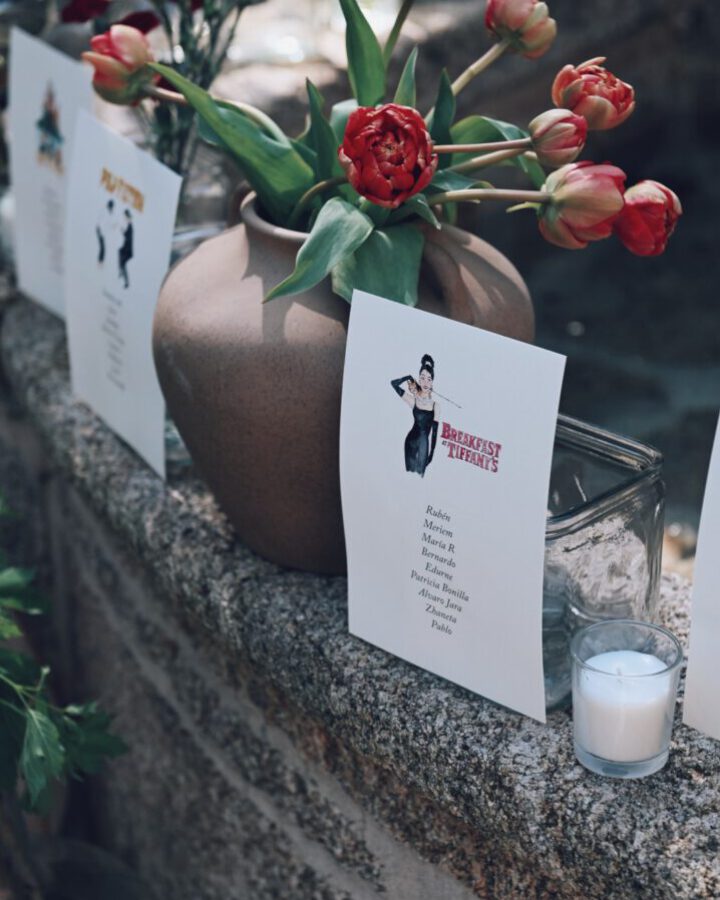 Tarjeta de seating plan con ilustración en acuarela de Breakfast at Tiffanys y lista de invitados ambientada con flores y vela