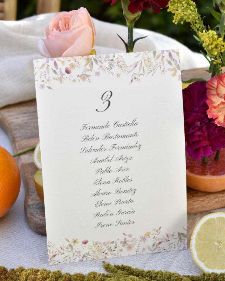 Seating plan de boda modelo Sabrina con flores rosadas en acuarela a mano ambientado sobre mesa con rosas rojas y decoración nupcial