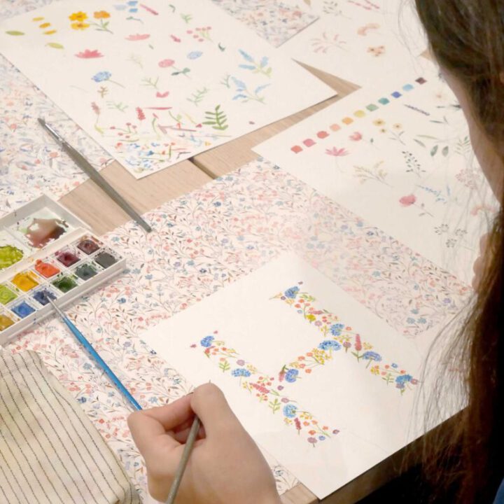 Manos de participante pintando estampado floral en acuarela con pincel fino sobre papel rodeado de ilustraciones botánicas