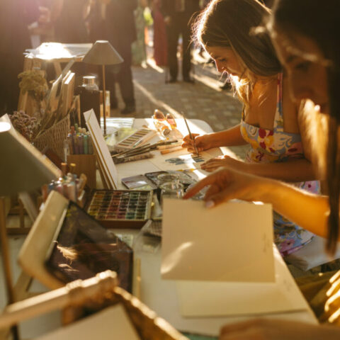 Artista pintando acuarelas en vivo durante celebración de boda al aire libre con invitados al fondo en jardín de Casa de Oficios