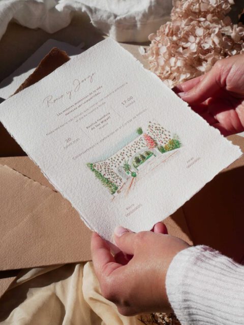 Manos sosteniendo una invitación de boda personalizada con ilustración del lugar de celebración pintada a mano en acuarela y tipografía caligráfica