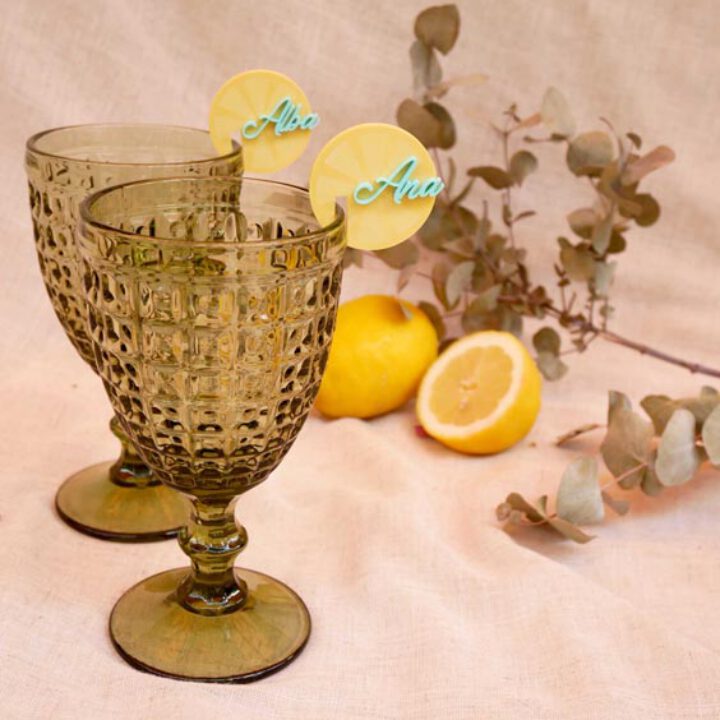 Marcacopas circulares amarillos con nombres en caligrafía colocados sobre copas de cristal tallado junto a limones y eucalipto
