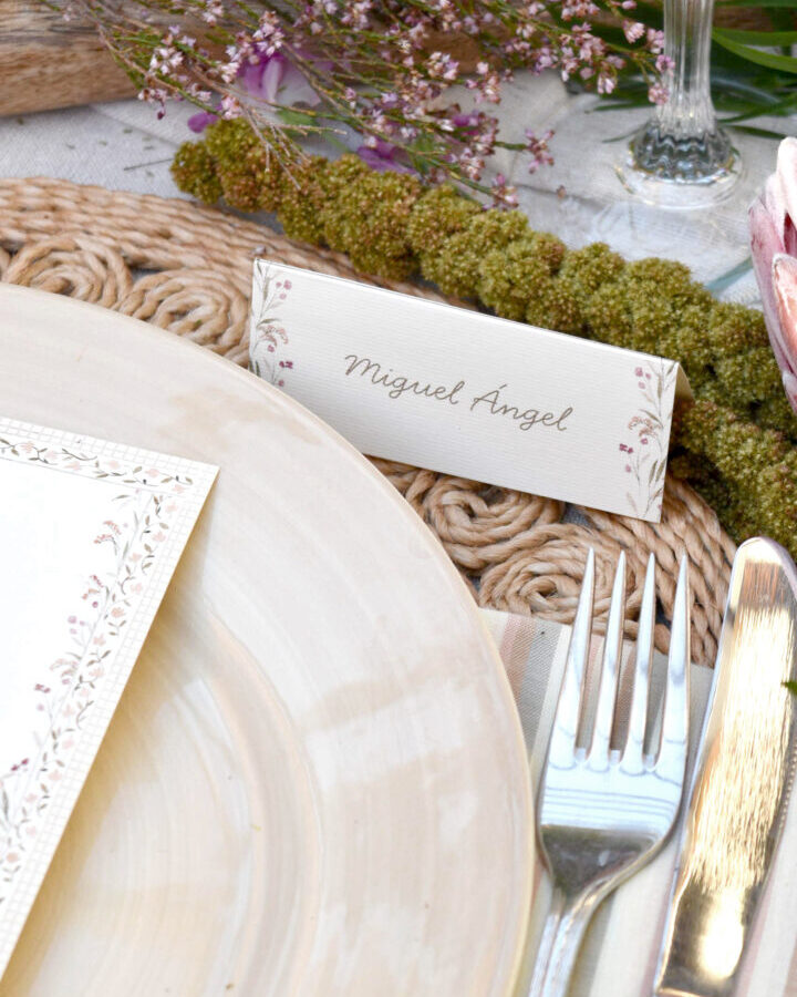 Marcasitio díptico vintage con ornamentos florales en acuarela y nombre caligráfico colocado sobre servilleta en mesa de boda rústica