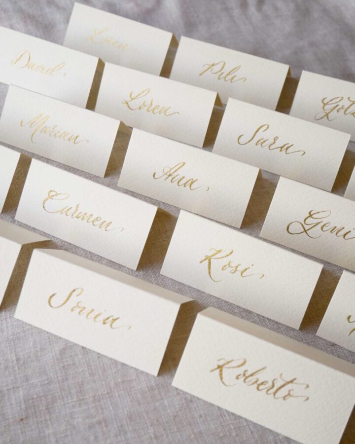 Marcasitios dípticos con nombres de invitados escritos en caligrafía artesanal con tinta dorada sobre papel color marfil