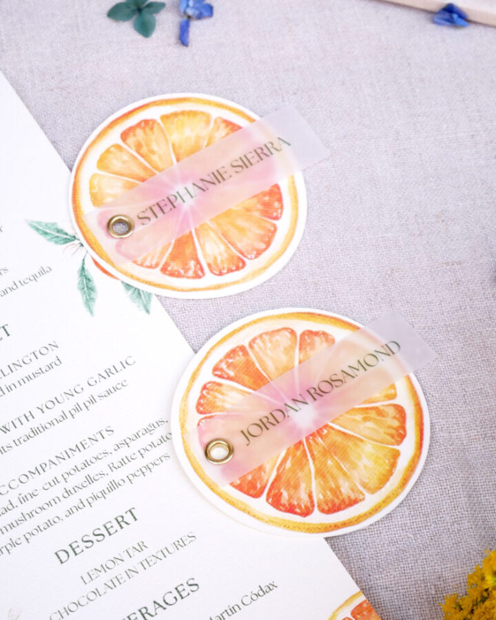 Dos marcasitios circulares con forma de naranja en acuarela colocados sobre una minuta de boda con hojas de eucalipto decorativas