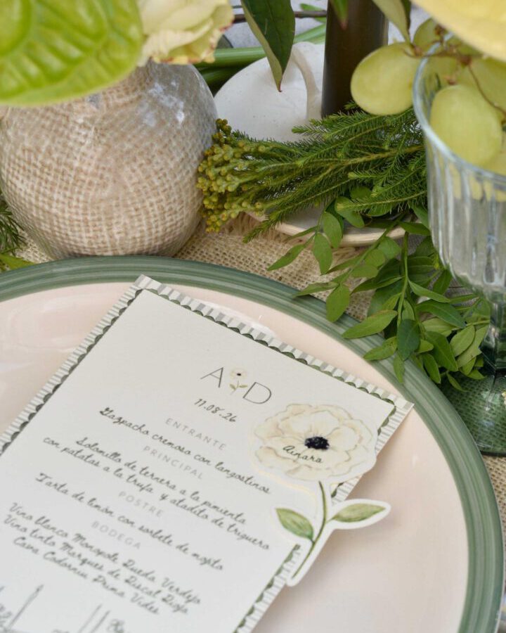 Marcasitio troquelado en forma de anémona con nombre del invitado junto a minuta caligráfica sobre plato de cristal con borde verde