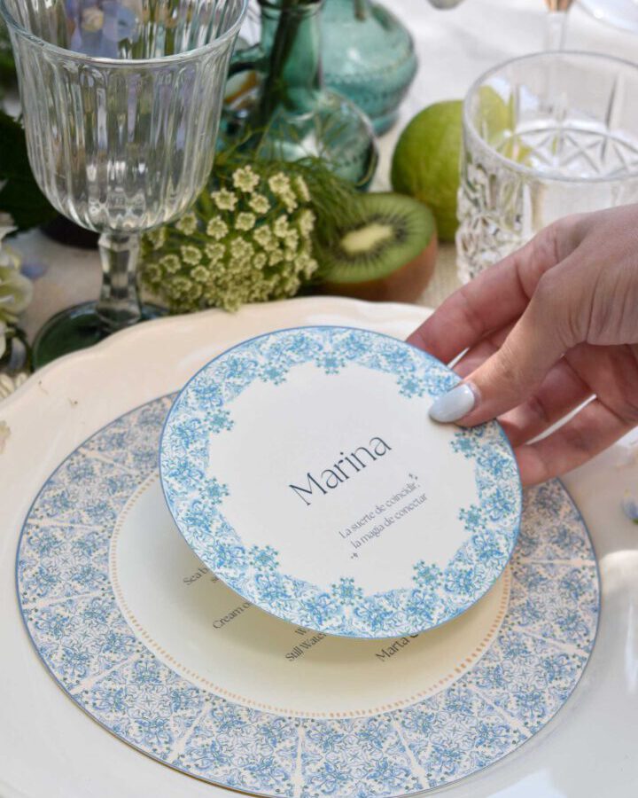 Marcasitio de estilo mediterráneo con nombre en caligrafía artesanal colocado sobre plato decorativo de cerámica azul en mesa de boda