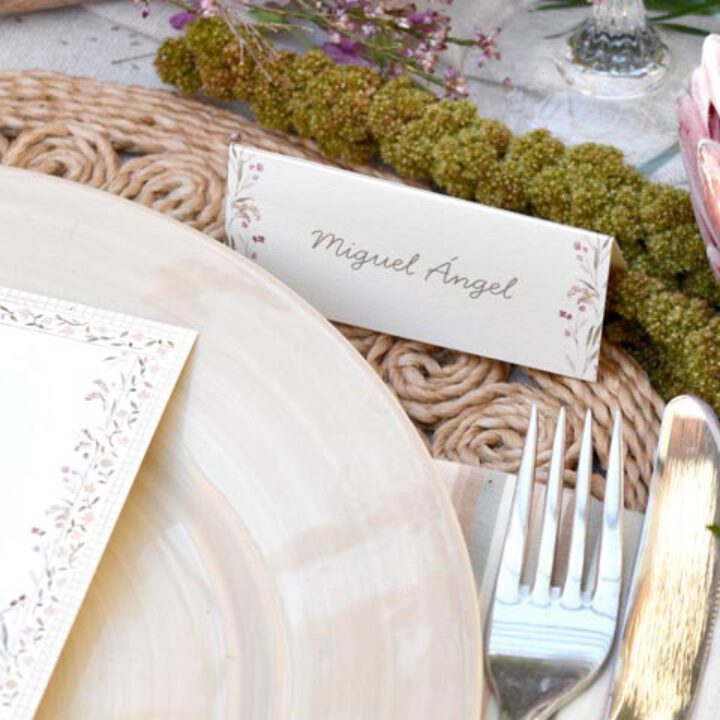 Marcasitios de boda con nombre en caligrafía y flores en acuarela sobre plato con cubertería dorada en mesa decorada con musgo
