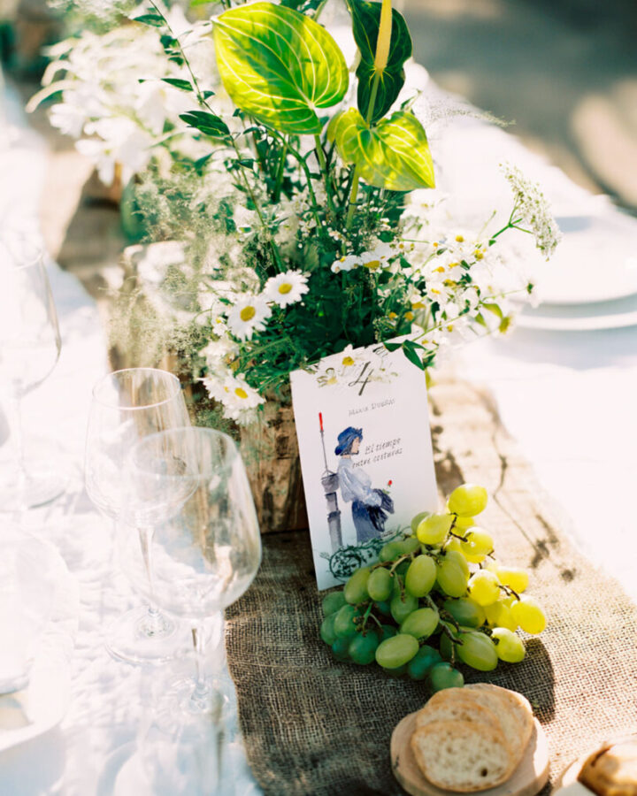 Mesero numerado con ilustración en acuarela de portada de libro sobre mesa de boda decorada con flores silvestres, uvas y copas
