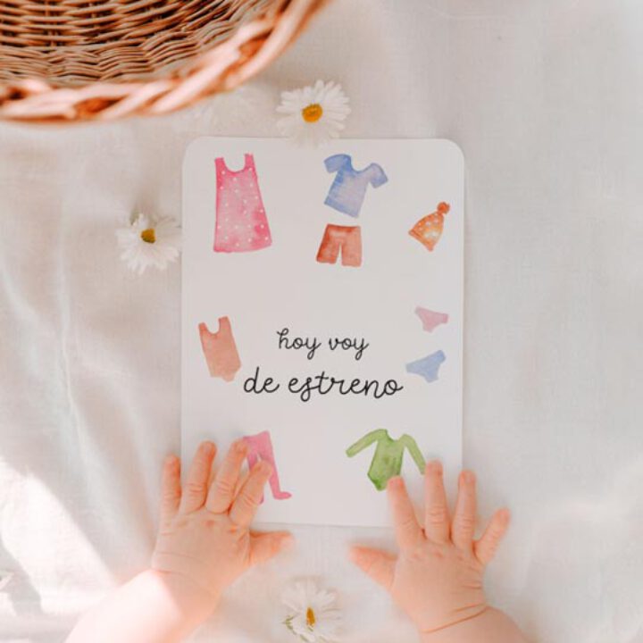 Manos de bebé sujetando una milestone card ilustrada con ropita en acuarela y texto hoy voy de estreno sobre tela blanca