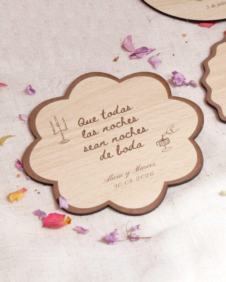 Posavasos de madera con forma de flor modelo Frida grabado con frase romántica, nombres y fecha sobre mesa con pétalos secos