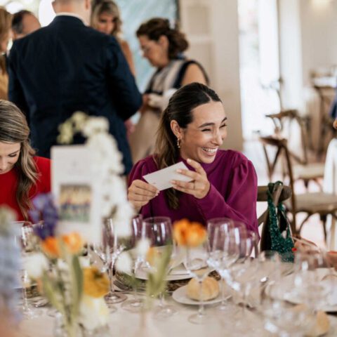 Invitada sonriente en mesa de banquete nupcial observando un detalle de papelería artesanal personalizado para la boda