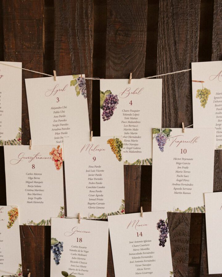 Seating plan de boda coleccion Uvas con tarjetas numeradas decoradas con racimos de uva acuarelados en tonos morados verdes y dorados colgadas en panel de madera rustico