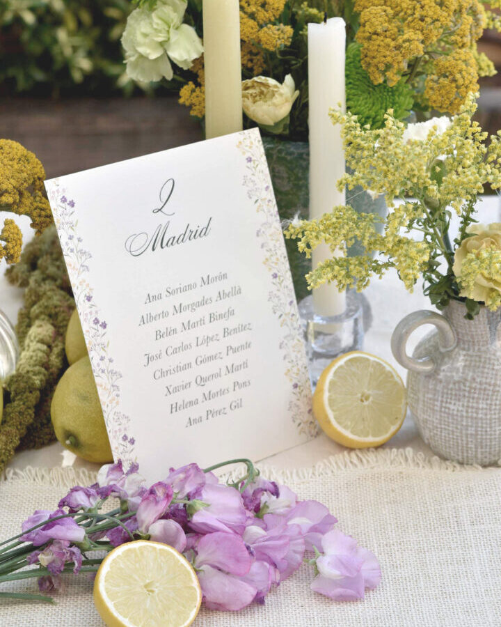 Tarjeta de seating plan de boda con nombre de mesa caligráfico, listado de invitados y cenefa decorativa de motivos florales en tonos lavanda