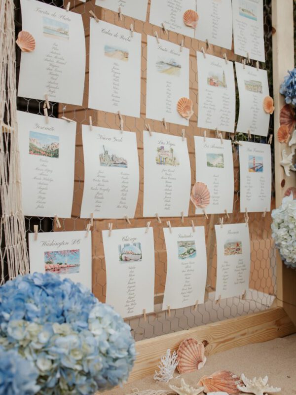 Seating plan de boda ilustrado con tarjetas individuales por mesa decoradas con acuarela artesanal y detalles marineros colgados en estructura decorativa