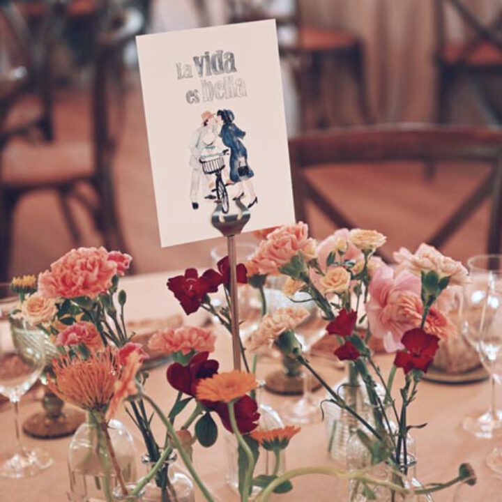 Tarjeta de seating plan ilustrada con dibujo de pareja y texto decorativo colocada entre arreglos florales en mesa de boda